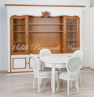 Tavolo rotondo in legno e sedie imbottite stile classico finitura colore bianco con libreria sullo sfondo